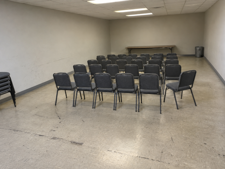 A plain rented meeting hall with grey stacking chairs in straight rows on a scuffed linoleum floor, bare walls and a folding table at the back, no people