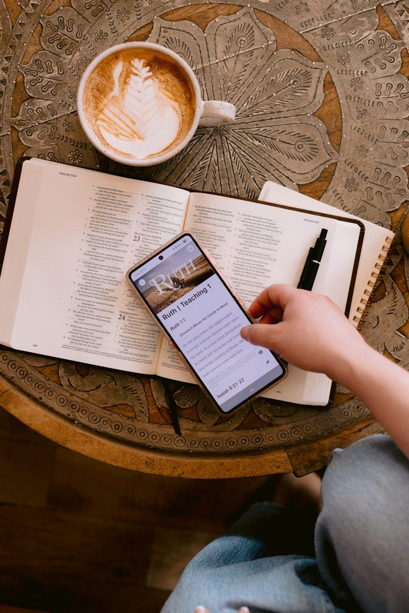 Overhead view of an open Bible, a notebook and pen, and a latte on a carved wooden table, with a hand holding a phone showing a Bible study app