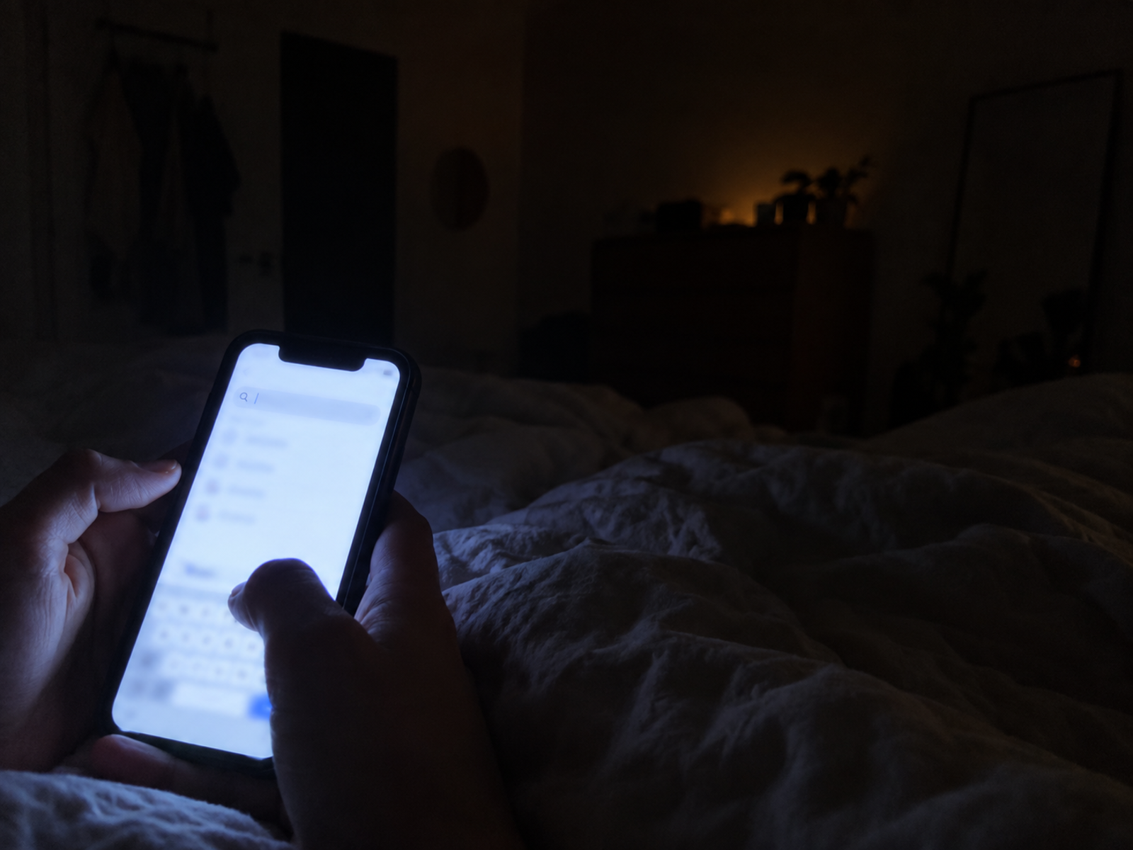 Over-the-shoulder view of hands holding a phone in a dark room at night, screen glow the only light, no face — a person searching at 2am